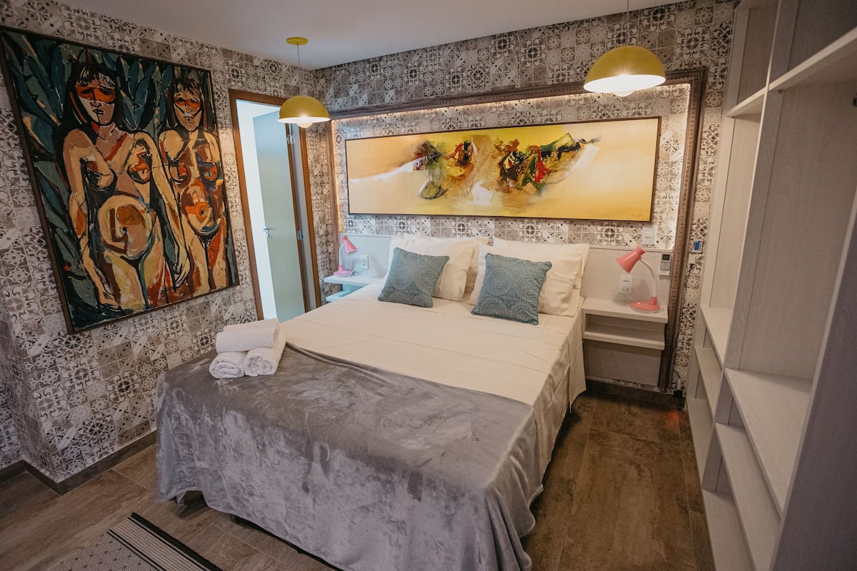 A bedroom with a large bed, two bedside tables, a painting on the wall, and patterned wallpaper.