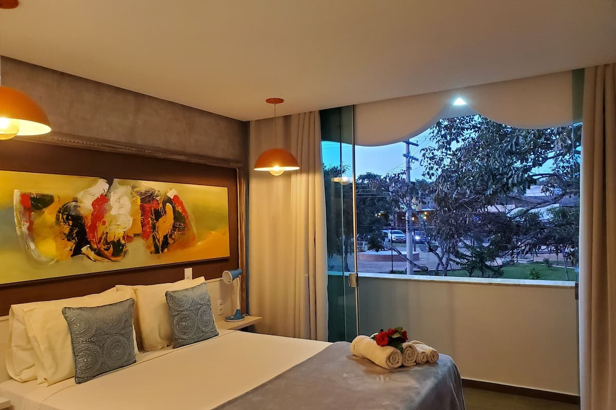 A hotel room with a large bed, a painting on the wall, a window with a view, and a bedside table with a lamp.
