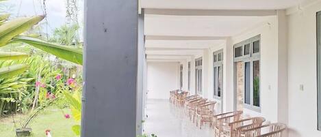 A covered outdoor area with rows of wooden chairs, a grassy garden, and a white building with large windows.