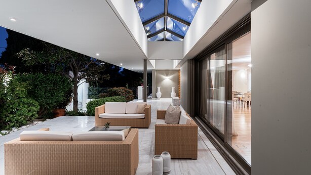 Un espacio exterior moderno con mobiliario de mimbre, techo de cristal y vistas al jardín.