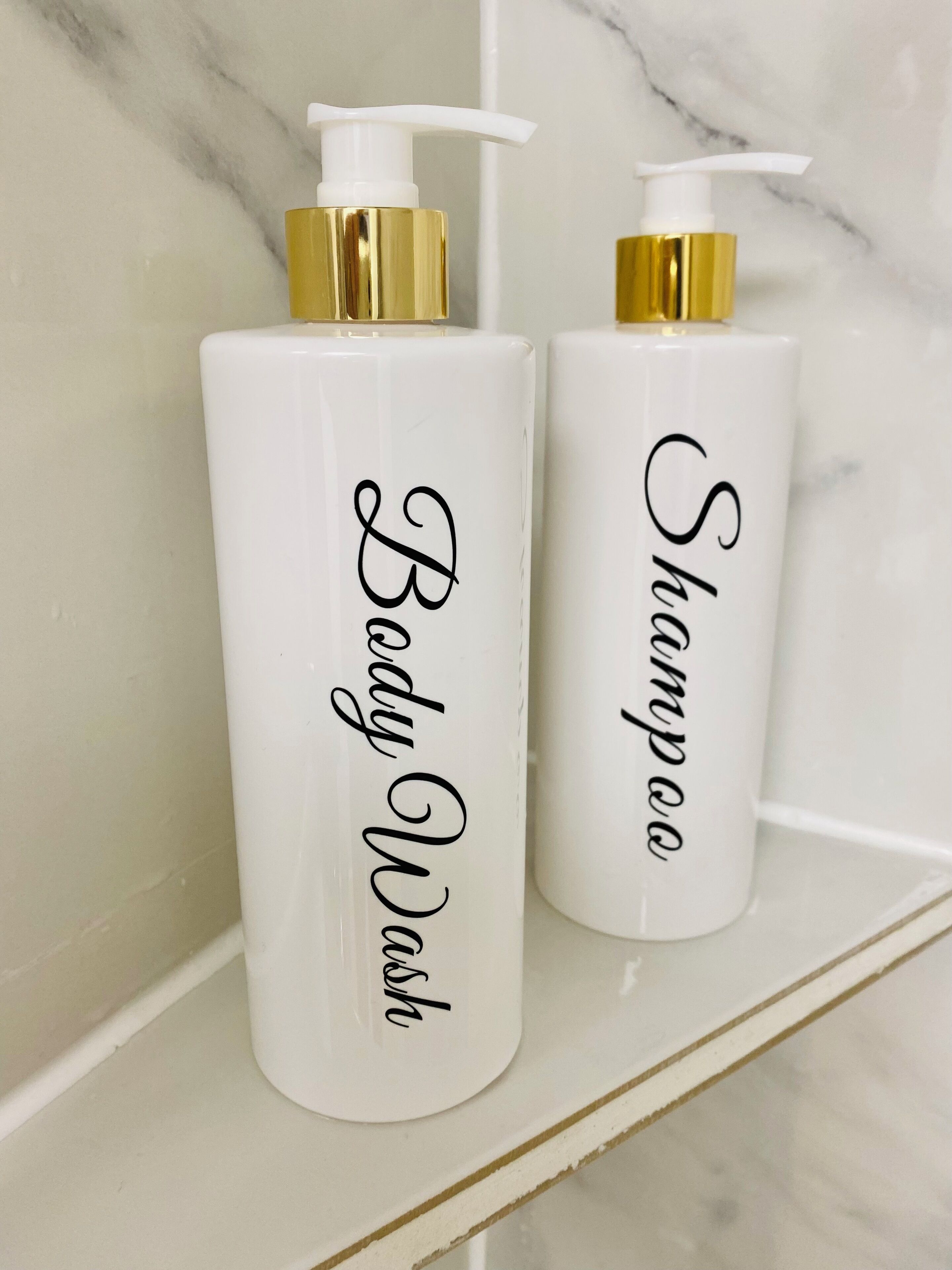 Two white bottles with gold pumps labeled "Body Wash" and "Shampoo" on a white shelf against a marble wall.