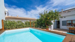 Une piscine dotée d’une terrasse en bois, entourée d’un bâtiment blanc avec un toit en tuiles et d’un jardin agrémenté d’un arbre.