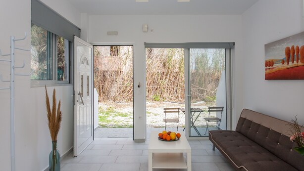 A modern living room with a sofa, a table with fruit, and a view through a glass door to an outdoor area. - Glaros Homes (Chania)