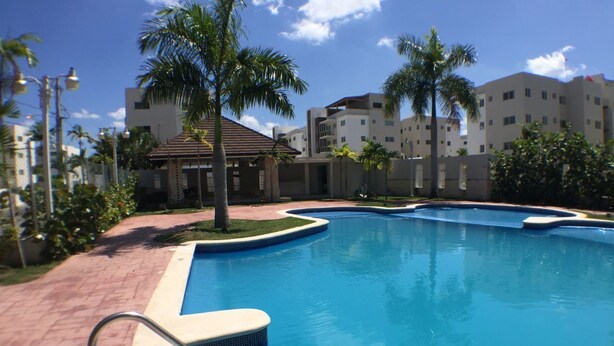 A swimming pool with a tiled deck, surrounded by palm trees and buildings. - GET AWAY Condo (Santiago De Los Caballeros)