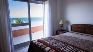 A bedroom with a bed, a nightstand, a lamp, and a view of the ocean through a sliding door. - Casa Milu - (Nazaré)