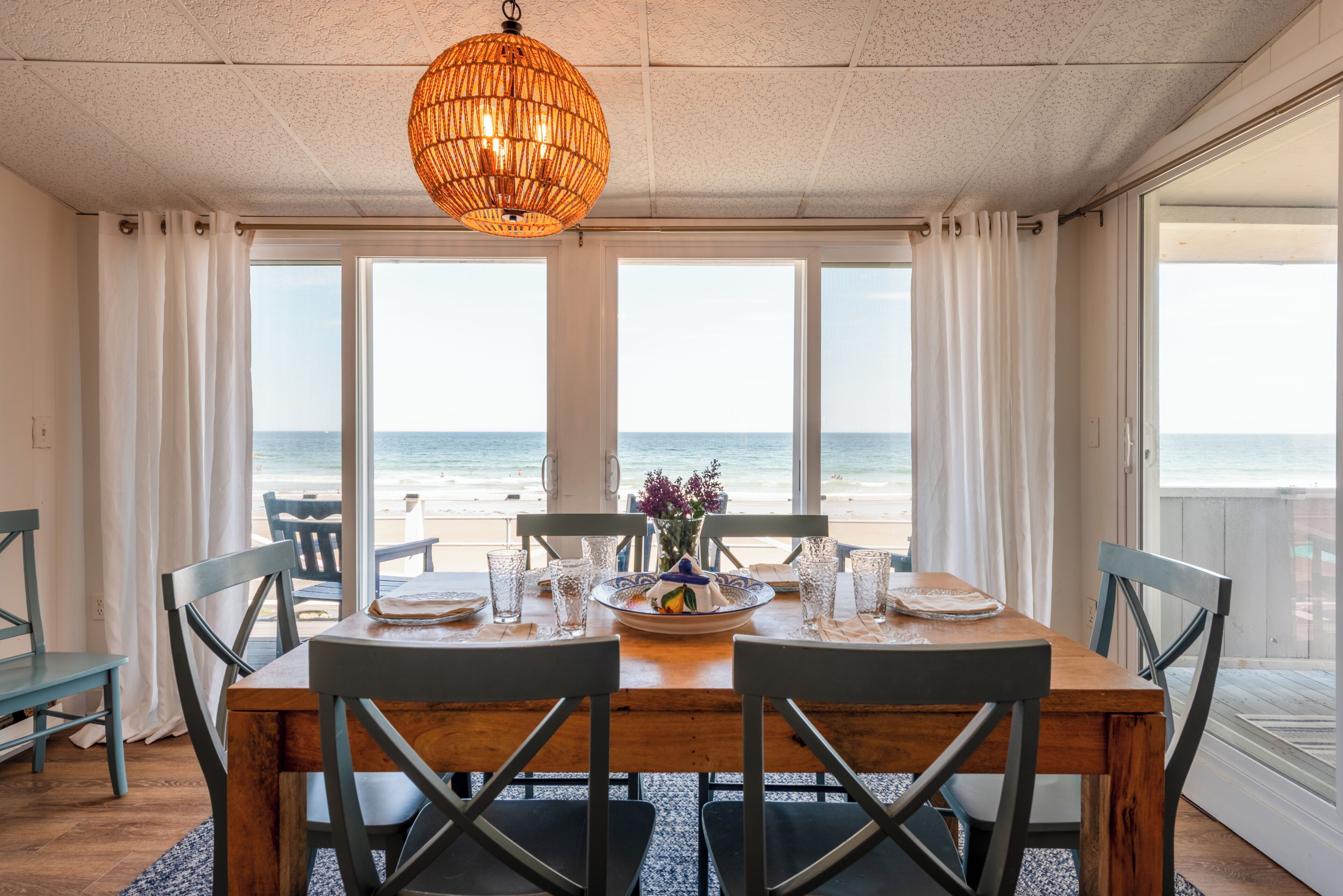 A dining area with a view of the beach, a hanging light fixture, and a table set for two.