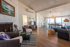 A spacious living room with a fireplace, a television, and a dining area with a view of the sea.