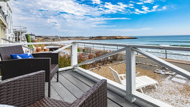 Una terraza de madera con mobiliario de mimbre y una silla blanca, con vistas a una playa de costa rocosa y un cielo azul despejado.