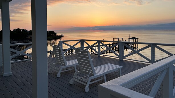 Une terrasse en bois, dotée de rampes blanches et de chaises longues, surplombe une étendue d’eau calme au coucher du soleil.