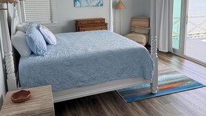 Une chambre avec un lit, un ventilateur de plafond, une fenêtre avec des stores, une table de chevet en bois, une lampe et une vue sur l’océan.