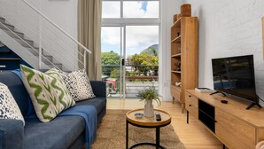 Een moderne woonkamer met een blauwe bank, een houten tv-meubel en een groot raam met uitzicht op groen en gebouwen.