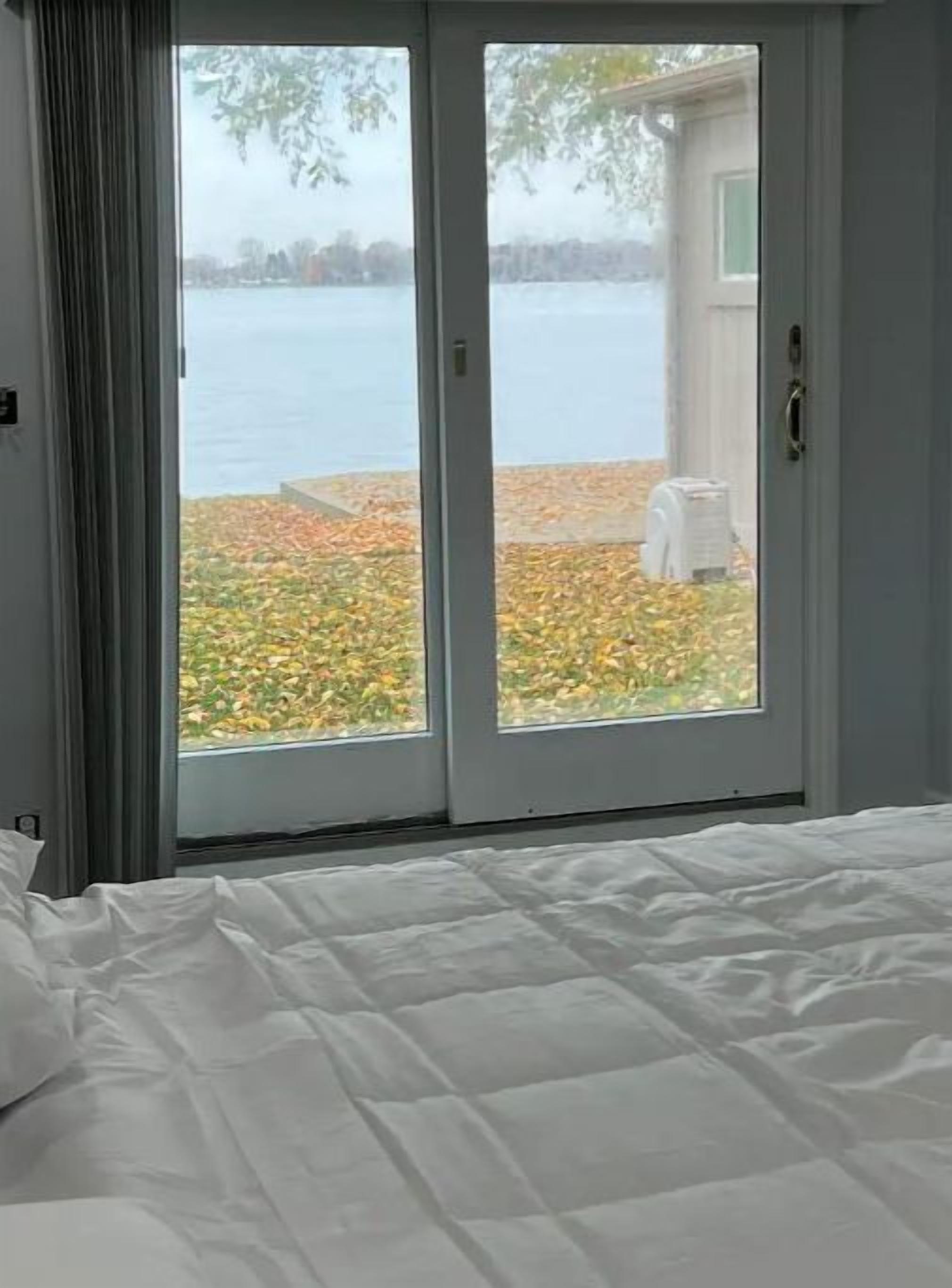 Un dormitorio con un ventanal grande y puerta corrediza de vidrio, con vistas a un lago y follaje otoñal.