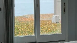 A bedroom with a large window and sliding glass door, offering a view of a lake and autumn foliage. - Cozy 3 bedroom fully furnished lake view house! (White Lake charter Township)