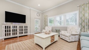 A living room with a television, a white cabinet, a coffee table, and a patterned chair. - Sunsets You'll Never Forget- Private Patio/Hot Tub/Heated Pool (Miramar Beach)