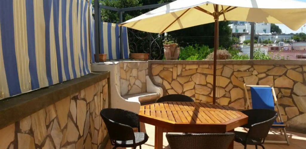 An outdoor patio with a stone wall, a wooden table, four chairs, and a large umbrella.