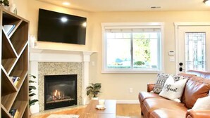 A living room with a fireplace, a television mounted above it, a leather sofa with a 'Guest' pillow, and a bookshelf.