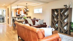 A spacious living room with a leather sofa, a wooden bookshelf, and a kitchen in the background.