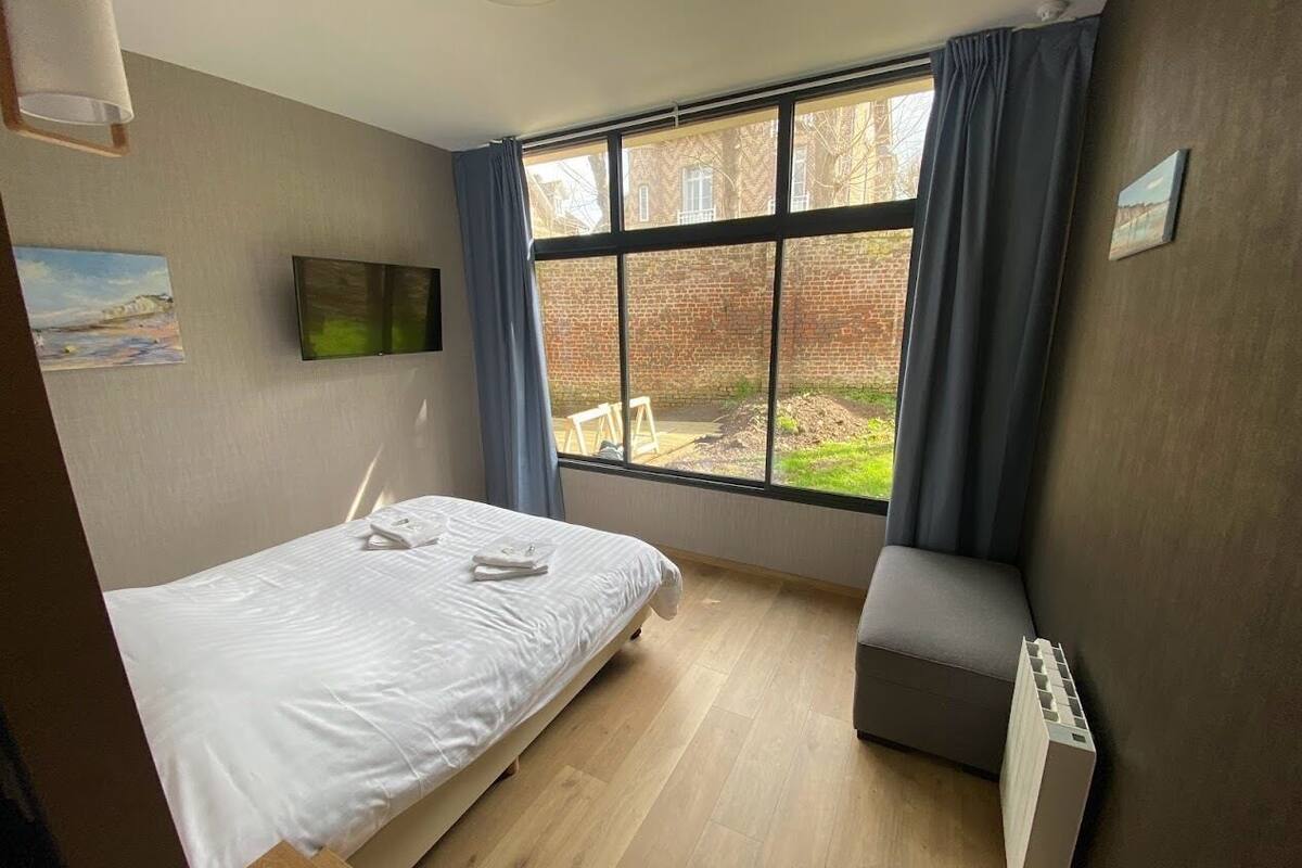 A hotel room with a bed, a small seating area, a window with curtains, and a TV mounted on the wall.