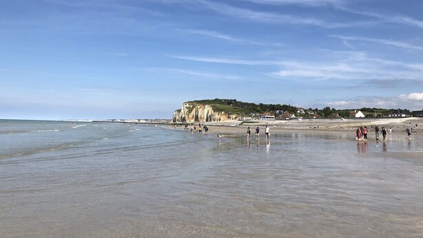 Una playa con un acantilado rocoso, cielo despejado y gente caminando por la orilla.