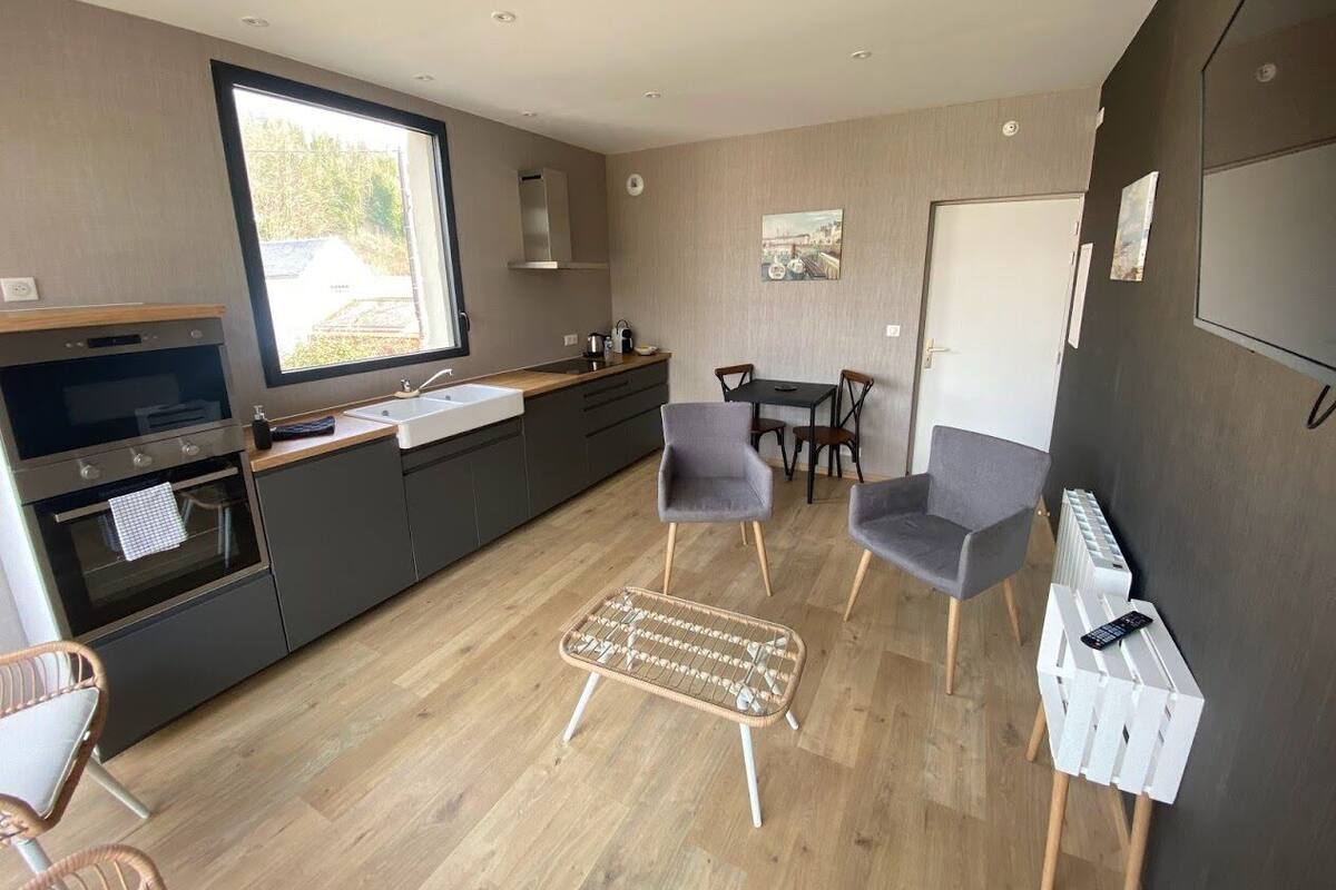 A modern kitchen with a large window, grey chairs, a small dining table, and a wicker ottoman.