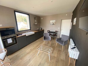 A modern kitchen with a large window, grey chairs, a small dining table, and a wicker ottoman. - Parc Beausejour Dieppe (Dieppe)