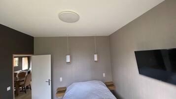 A bedroom with a bed, bedside tables, a TV mounted on the wall, and a door leading to a dining area.