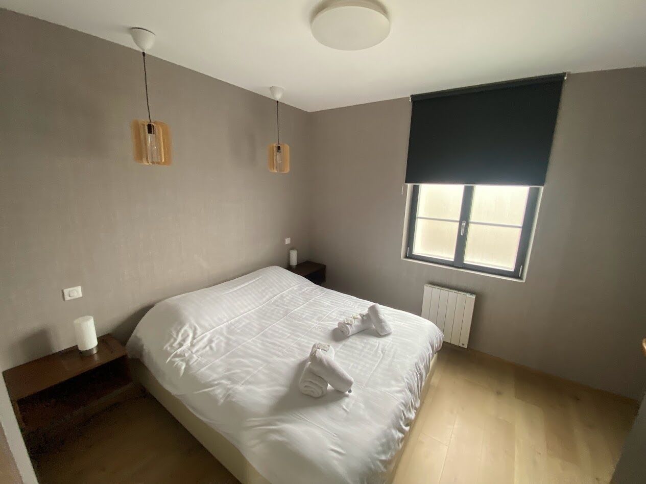 A bedroom with a bed, two bedside lamps, a window with blinds, and a radiator.