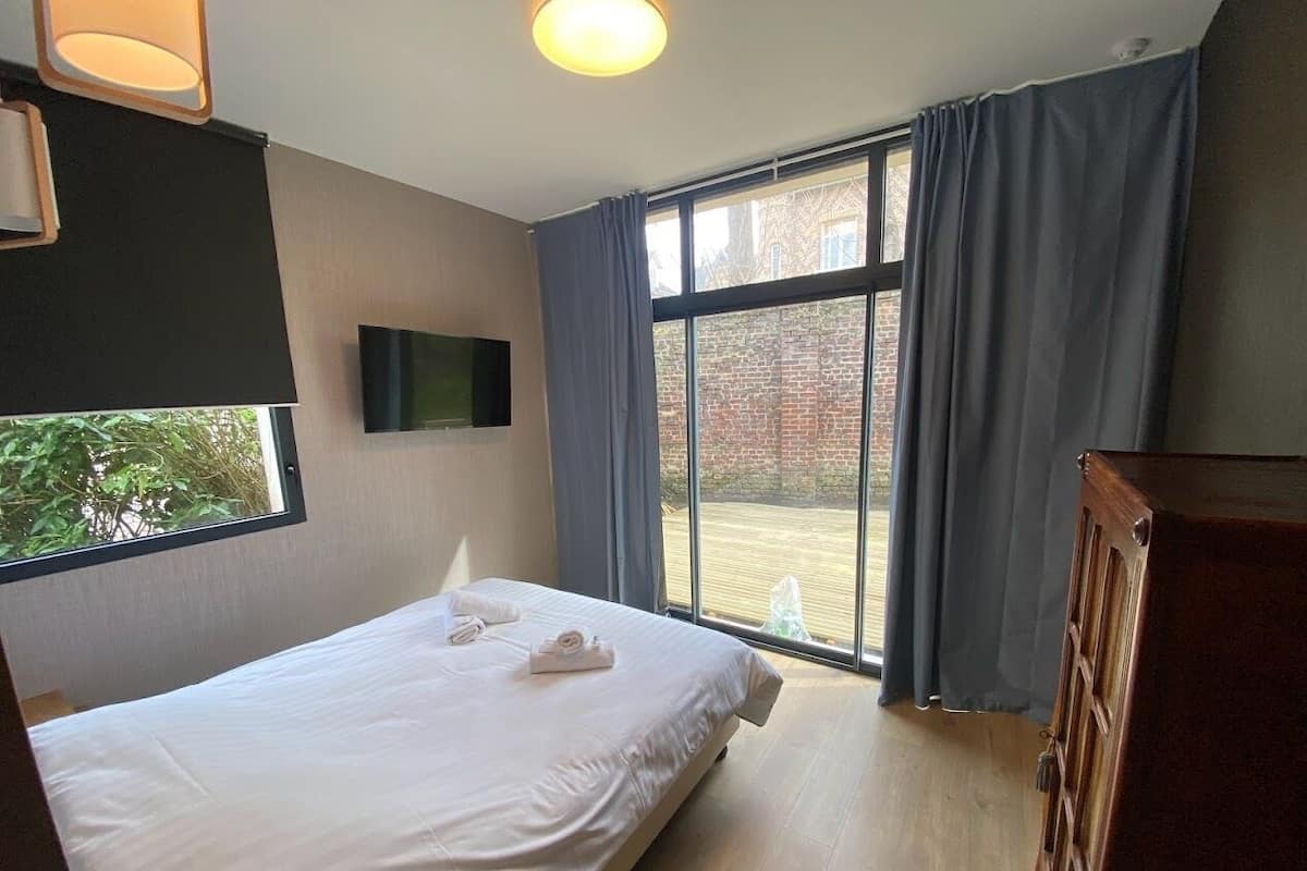 A hotel room with a bed, a TV, a window with curtains, and a view of the outside.