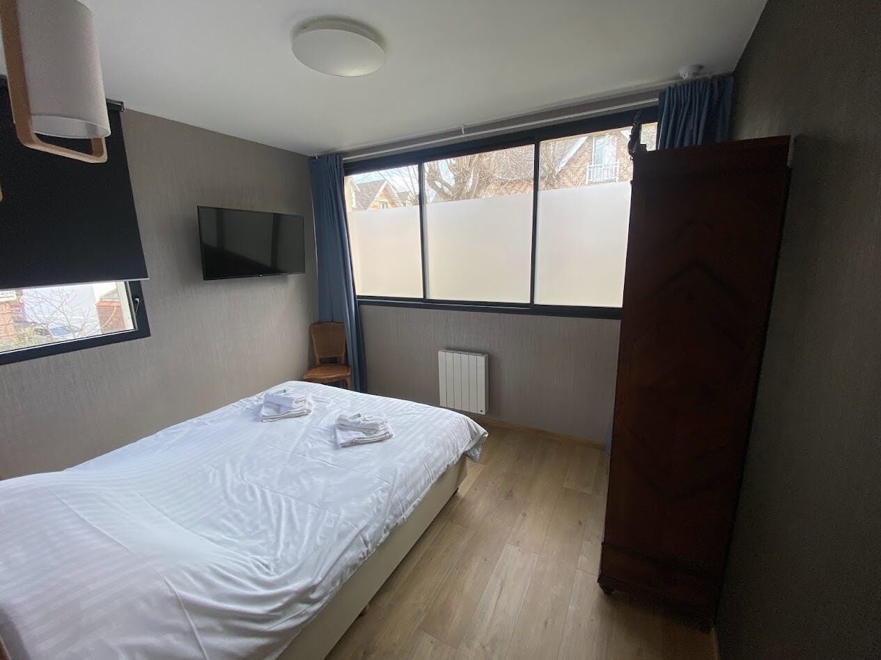 Un lit simple avec du linge de lit blanc, un téléviseur à écran plat fixé au mur, une fenêtre avec des stores et un radiateur.