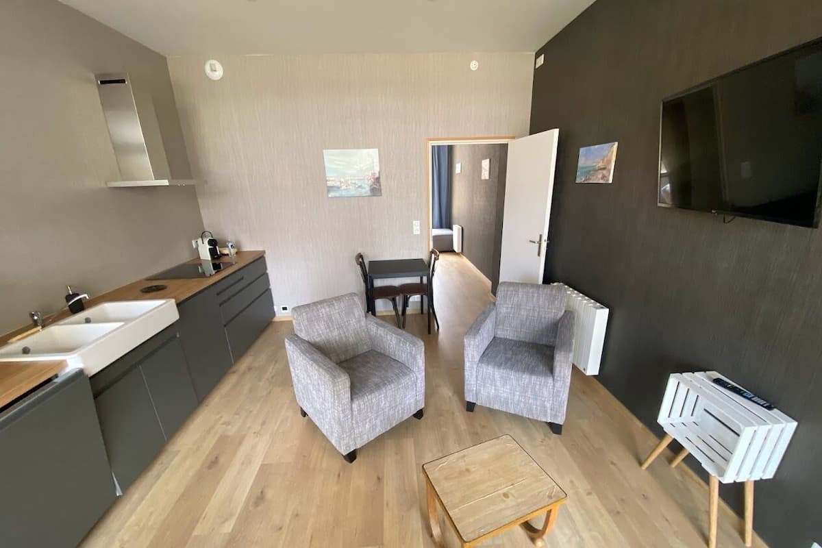A modern living room with a kitchen area, two grey armchairs, a wooden coffee table, and a TV mounted on the wall.
