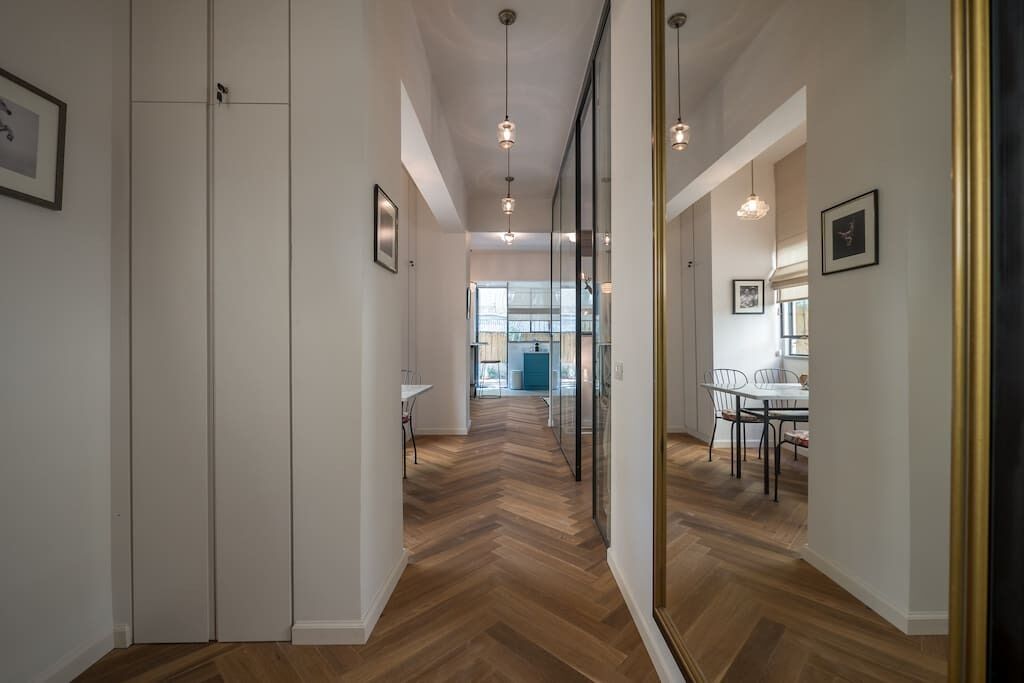 Un couloir doté d’un parquet en chevrons, de murs blancs et de tableaux encadrés. On aperçoit un coin repas avec une table et des chaises à travers une cloison en verre.