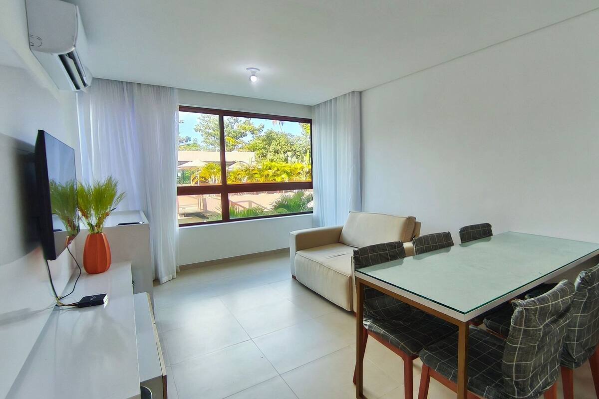Una sala de estar moderna con una mesa de comedor de vidrio, un sofá, un televisor y un ventanal con vista a zonas verdes.