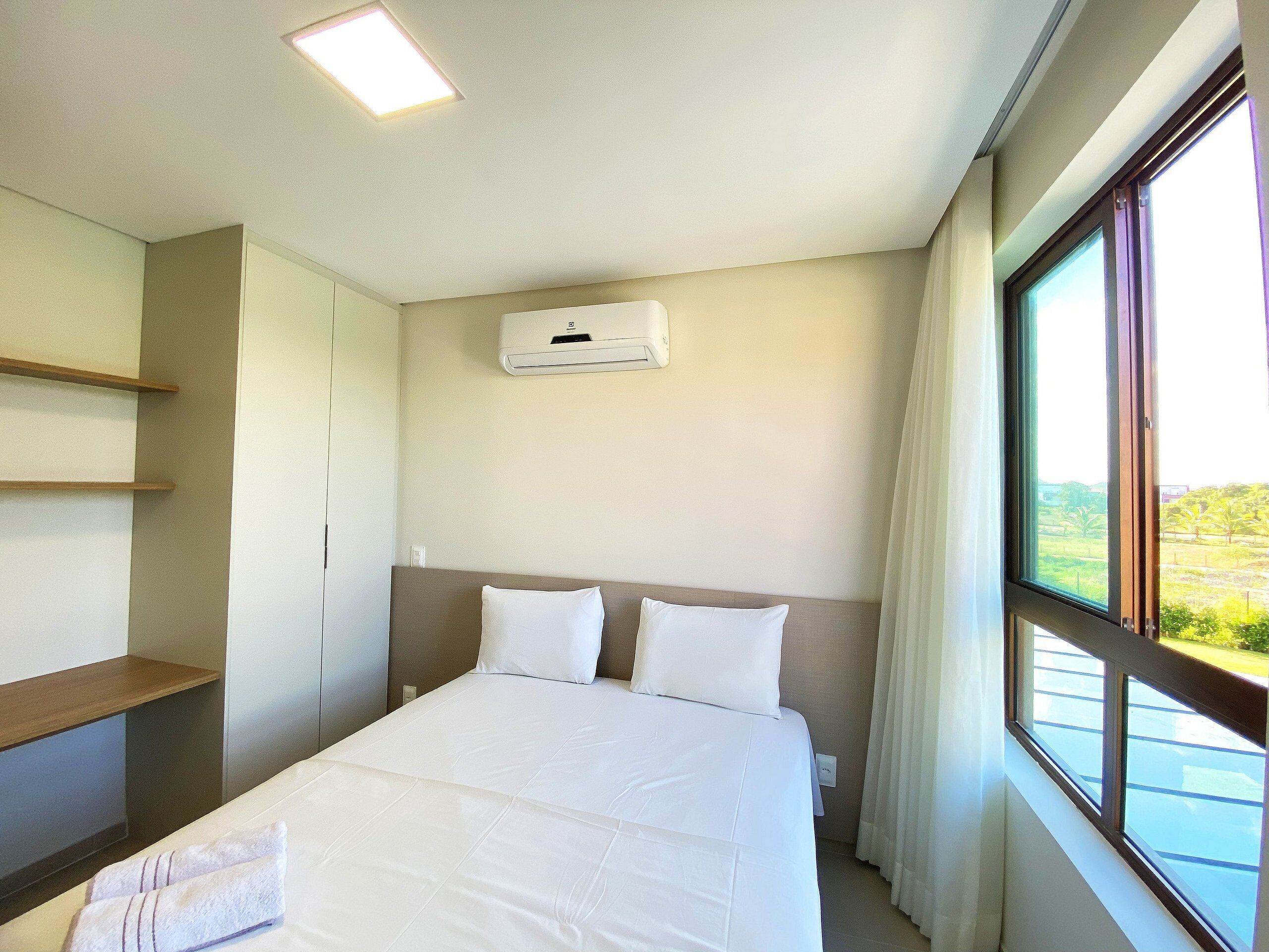 Een hotelkamer met een bed, een raam met gordijnen, een wandairconditioner en een houten plank.
