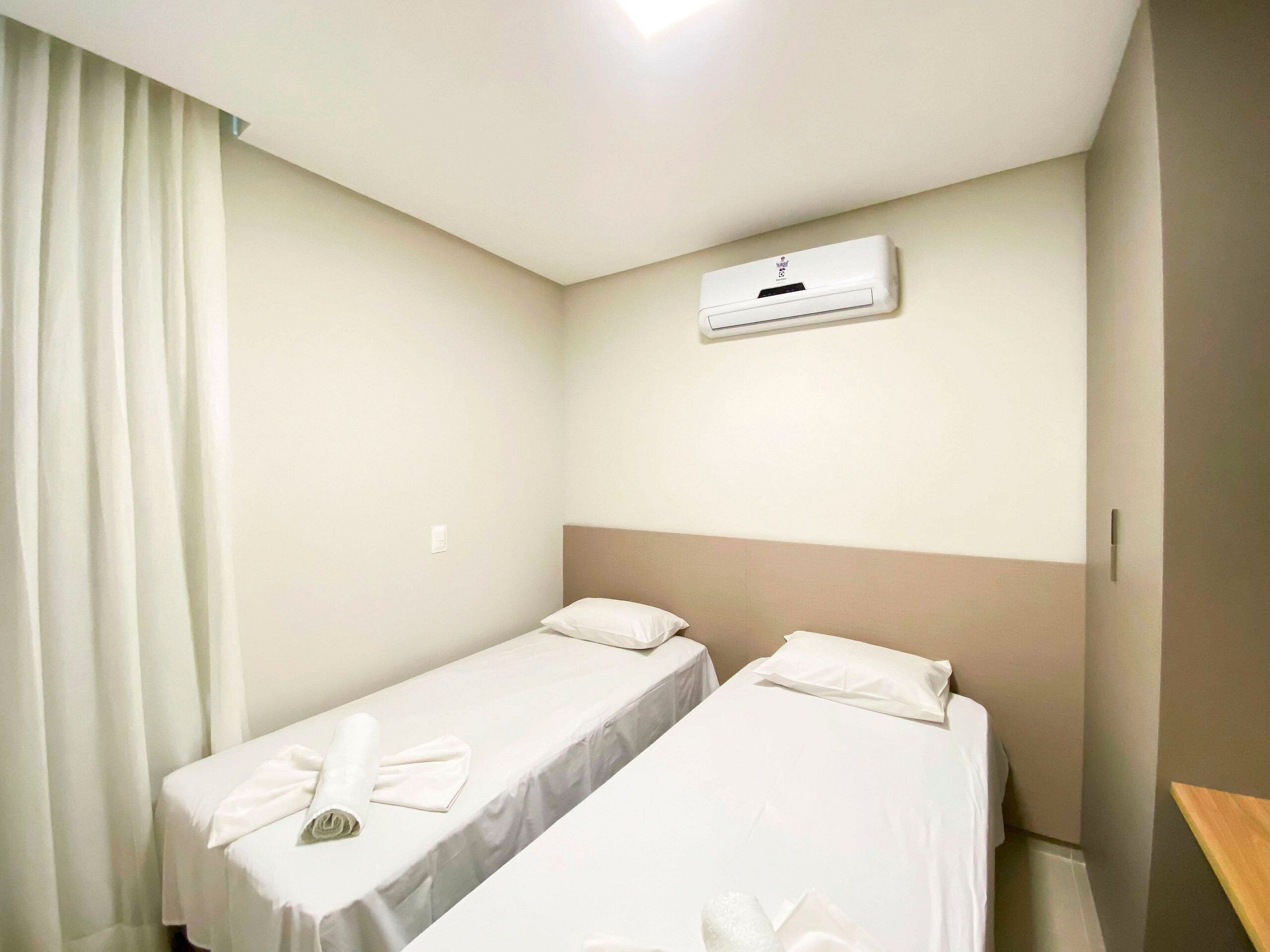 Quarto de hotel com duas camas de solteiro, ar condicionado no teto e janela com cortinas.