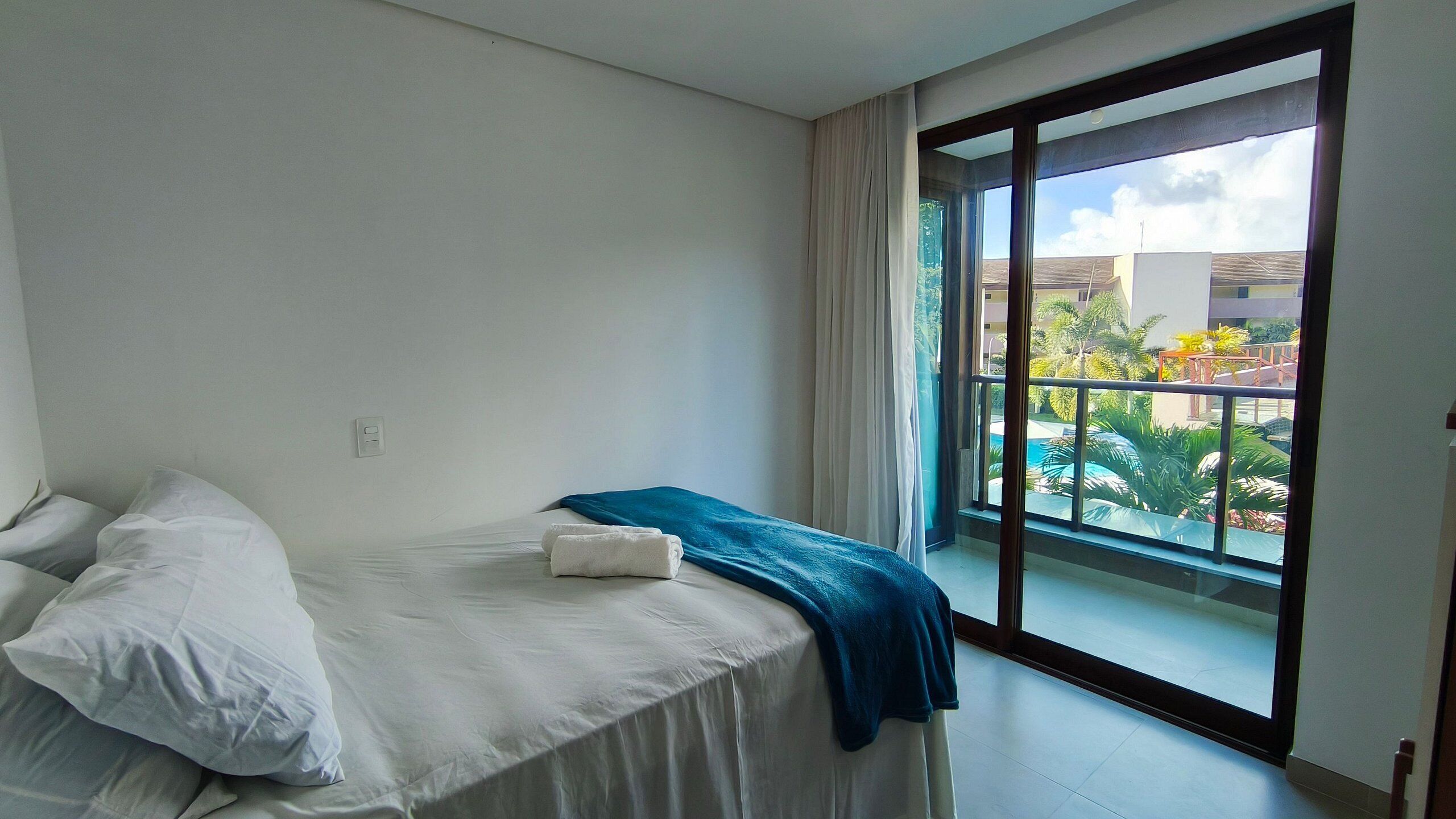 Quarto de hotel com cama, janela e vista para um prédio e vegetação lá fora.