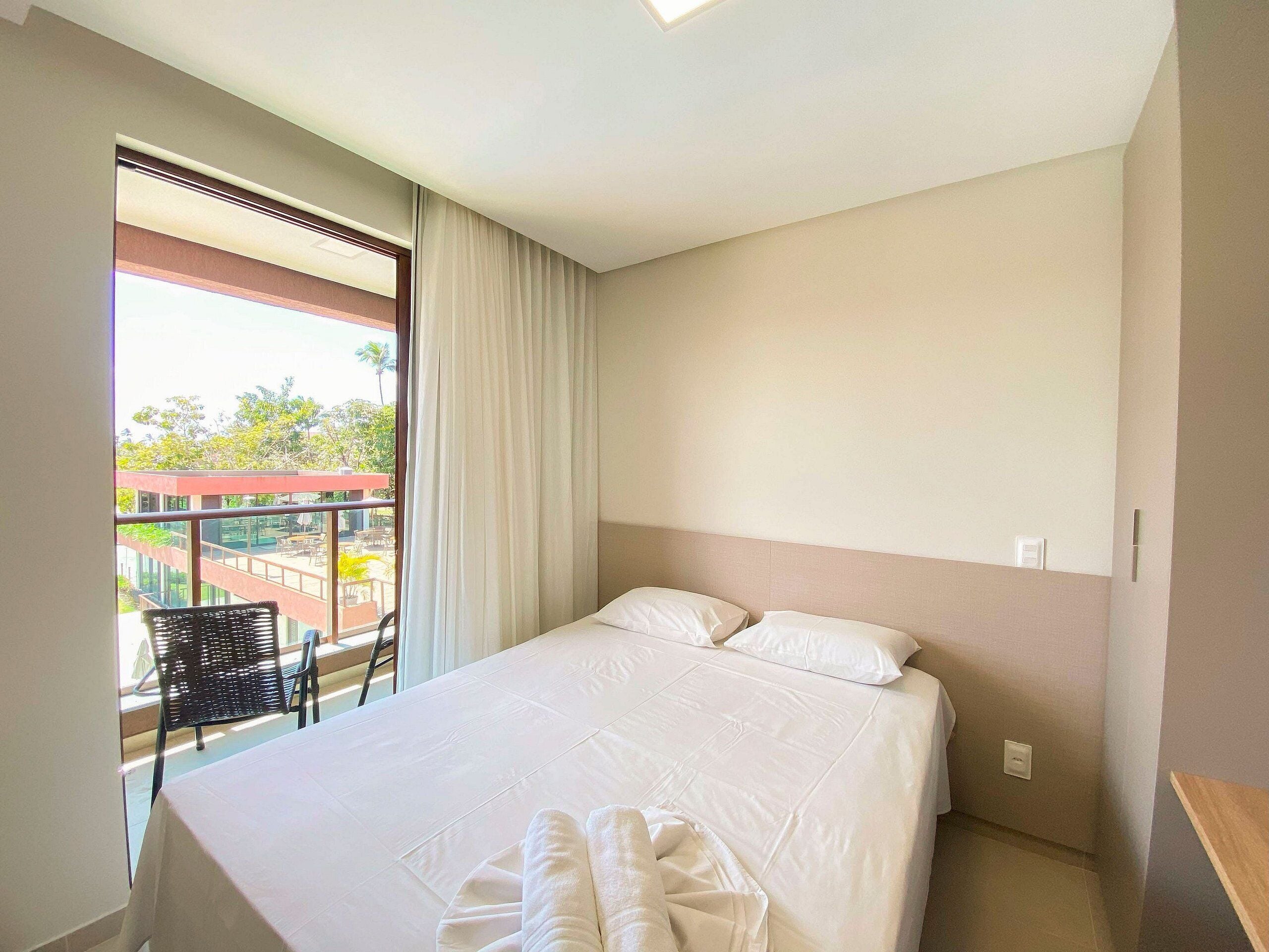 Quarto de hotel com cama, janela com cortinas, varanda com cadeira e vista para o exterior.