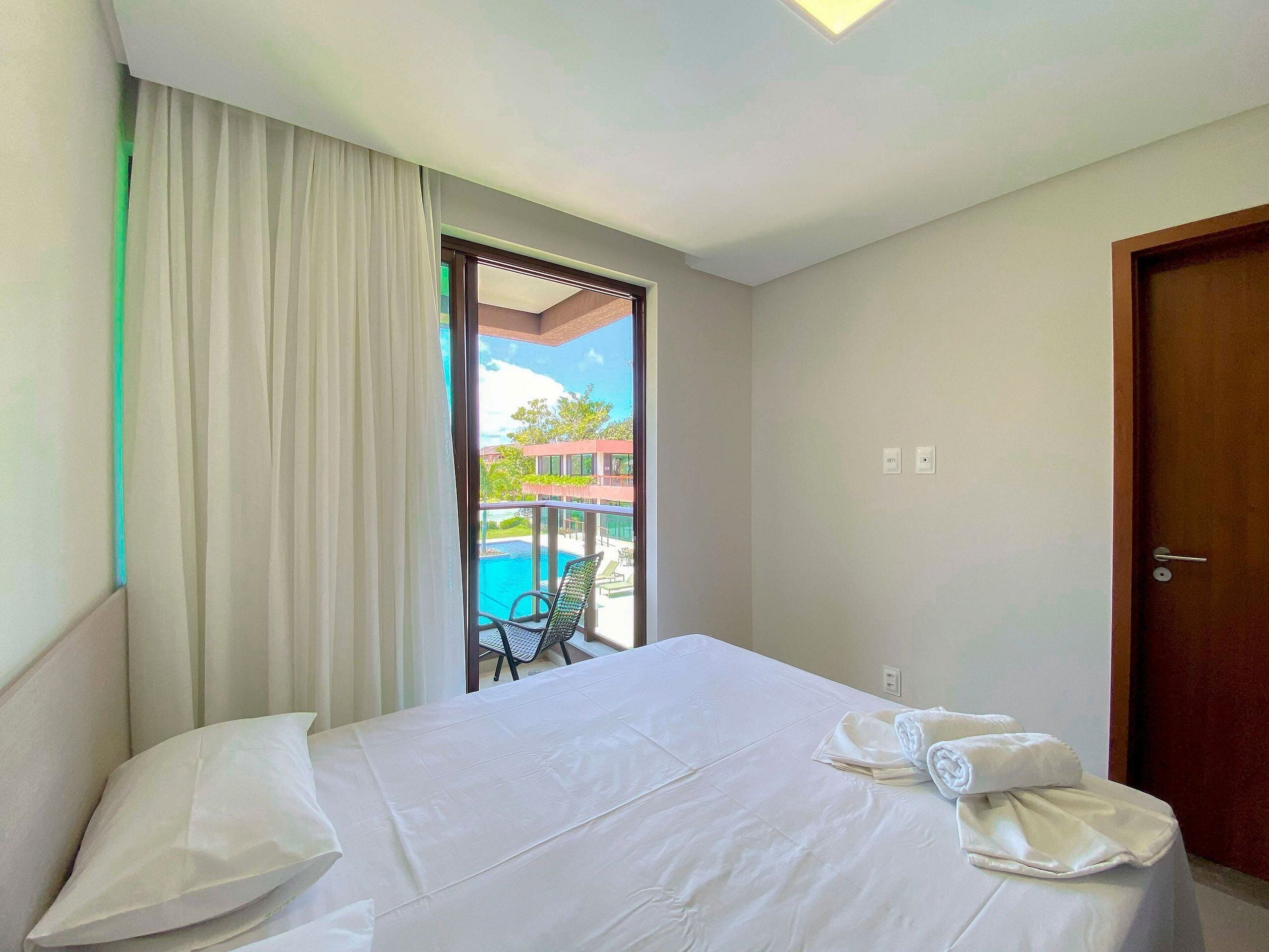 Quarto de hotel com cama, janela com cortinas, porta e vista para uma piscina e um edifício.