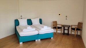 A double bed with white linens and a teal headboard, two wooden chairs, a small round table, and a wall socket. - Hotel Lethehof (Wardenburg)