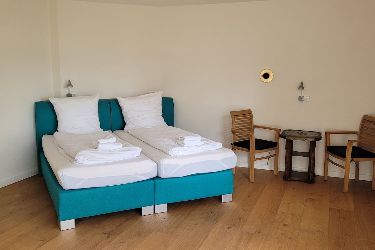 A double bed with white linens and a teal headboard, two wooden chairs, a small round table, and a wall socket.