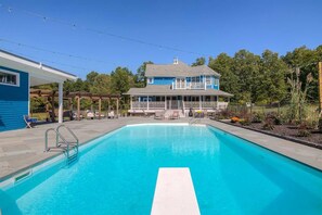 Une piscine bordée de blanc, un patio couvert et une maison à l’extérieur bleue, avec une balustrade blanche sur le porche.
