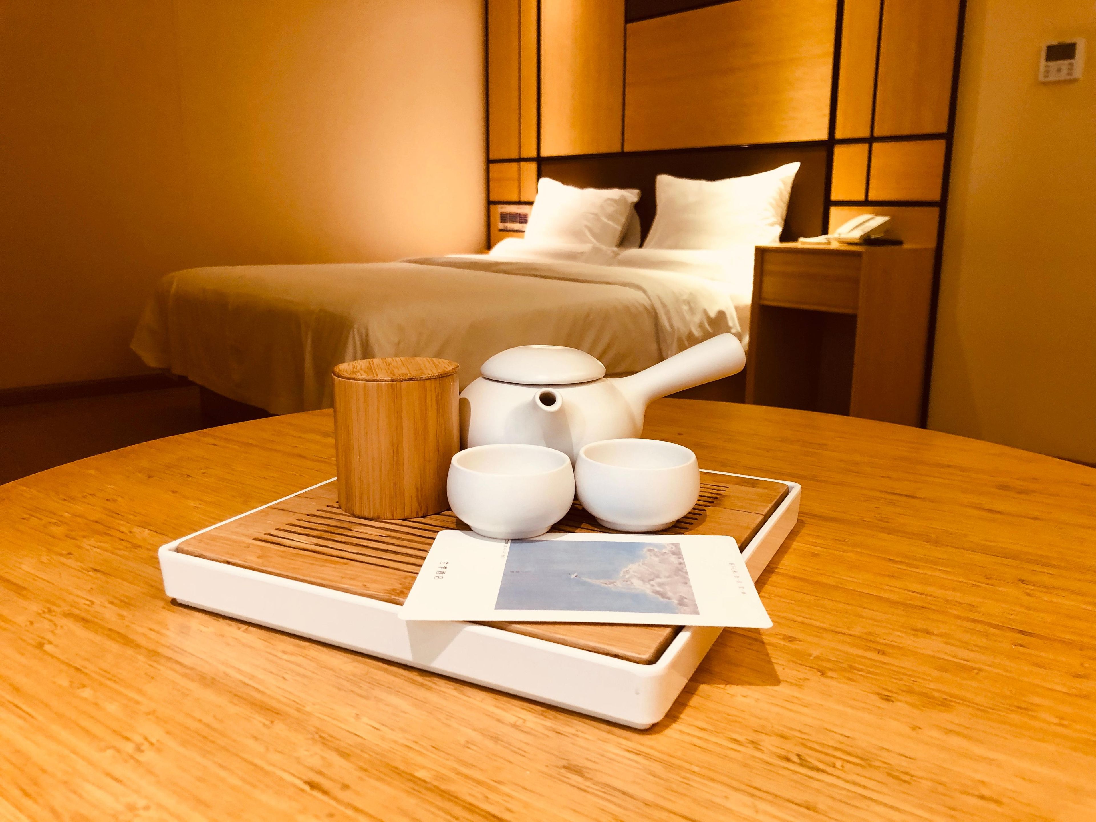 A neatly arranged hotel room with a bed, bedside table, and a table with a teapot, cups, and a tray with a photo.
