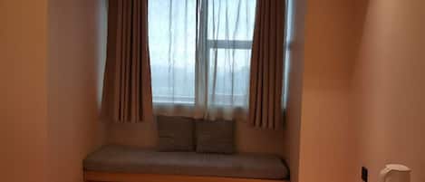 Una habitación con un banco integrado, una ventana con cortinas, una pequeña mesa con una taza blanca y un enchufe de pared.
