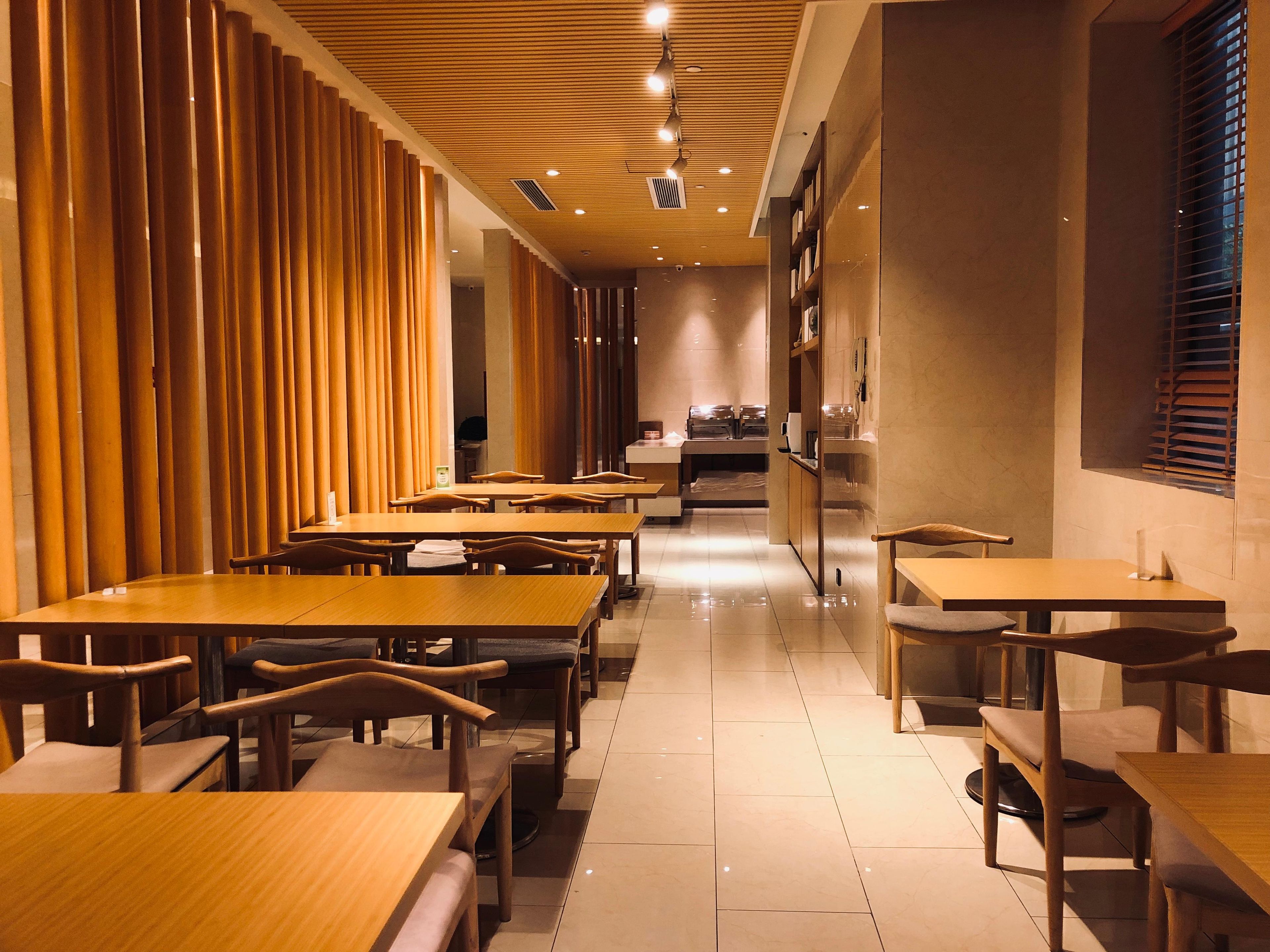 A modern interior with wooden tables and chairs, a kitchen area in the background, and a ceiling with recessed lighting.