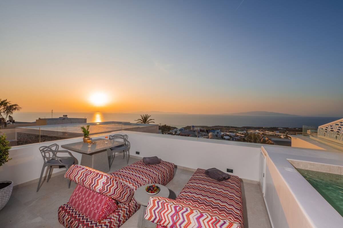 Una terraza en la azotea con una mesa y sillas, un conjunto de sofás y vistas a la puesta de sol sobre el mar.