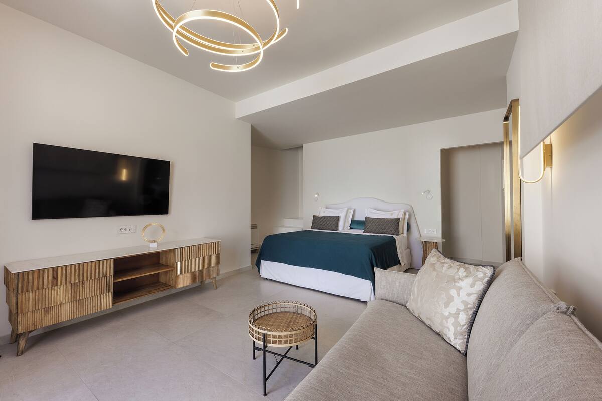 Un dormitorio moderno con una cama grande, un televisor montado en un mueble de madera, una mesita redonda de mimbre y un sofá.