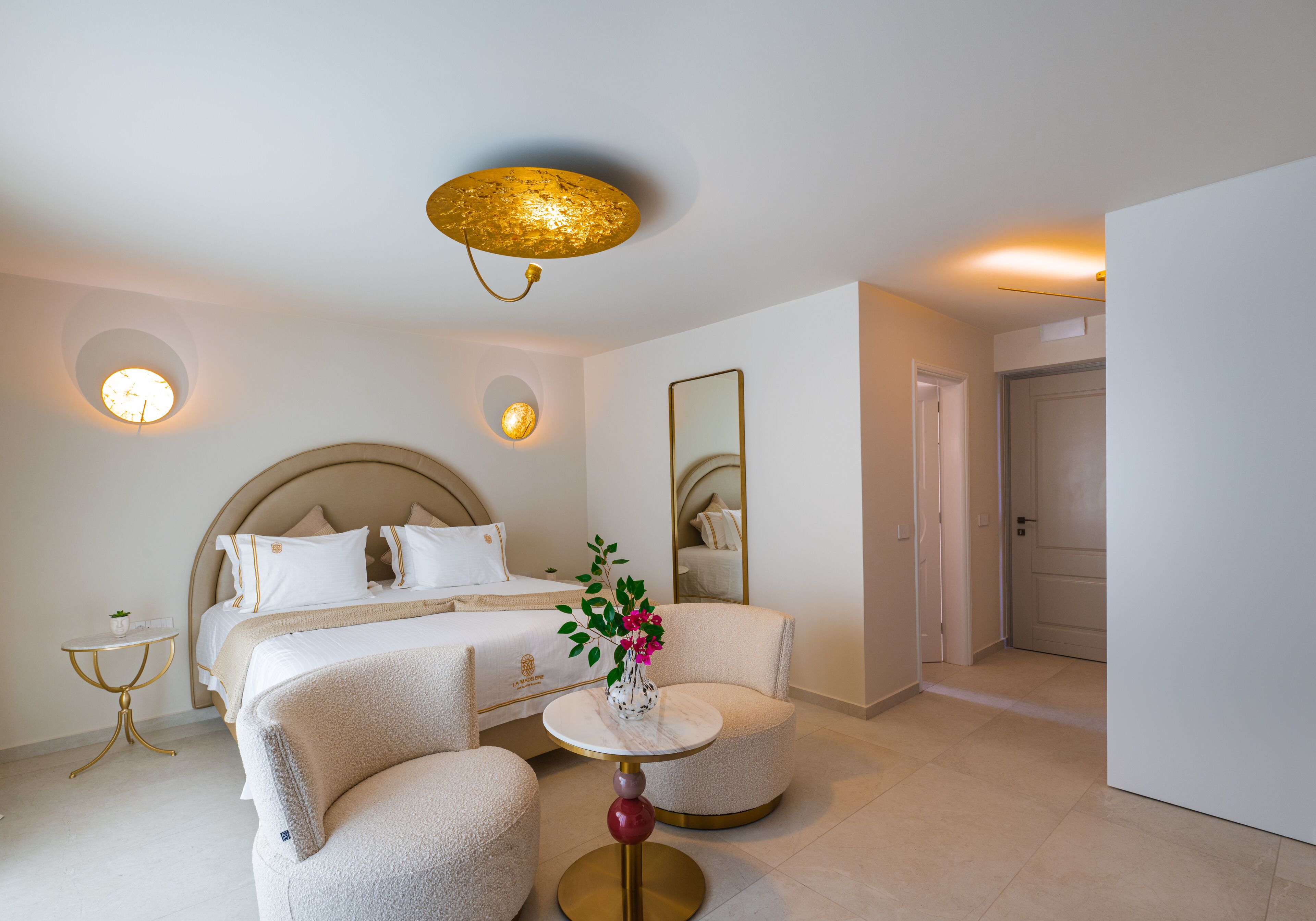 Una habitación de hotel con una cama, dos sillones, una mesita redonda con un florero y una lámpara de techo redonda, además de un espejo en la pared.