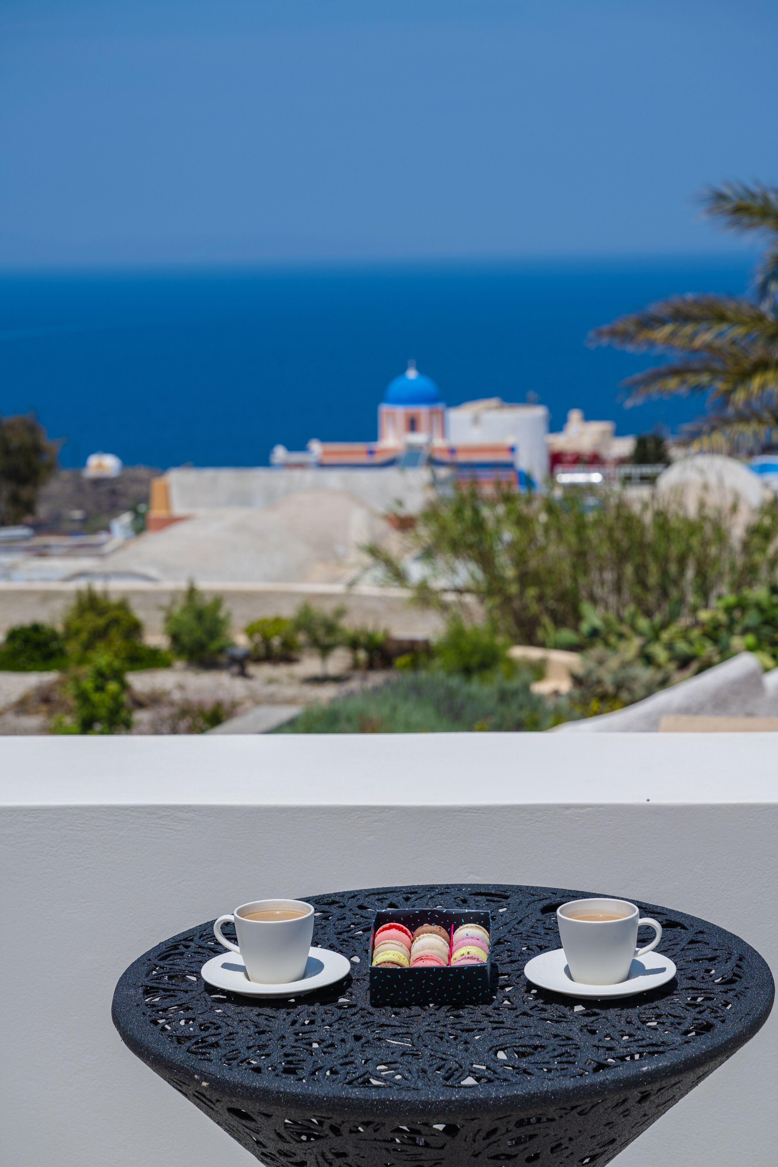 A table with two cups of coffee and a box of macarons, overlooking a coastal town with buildings and the sea.