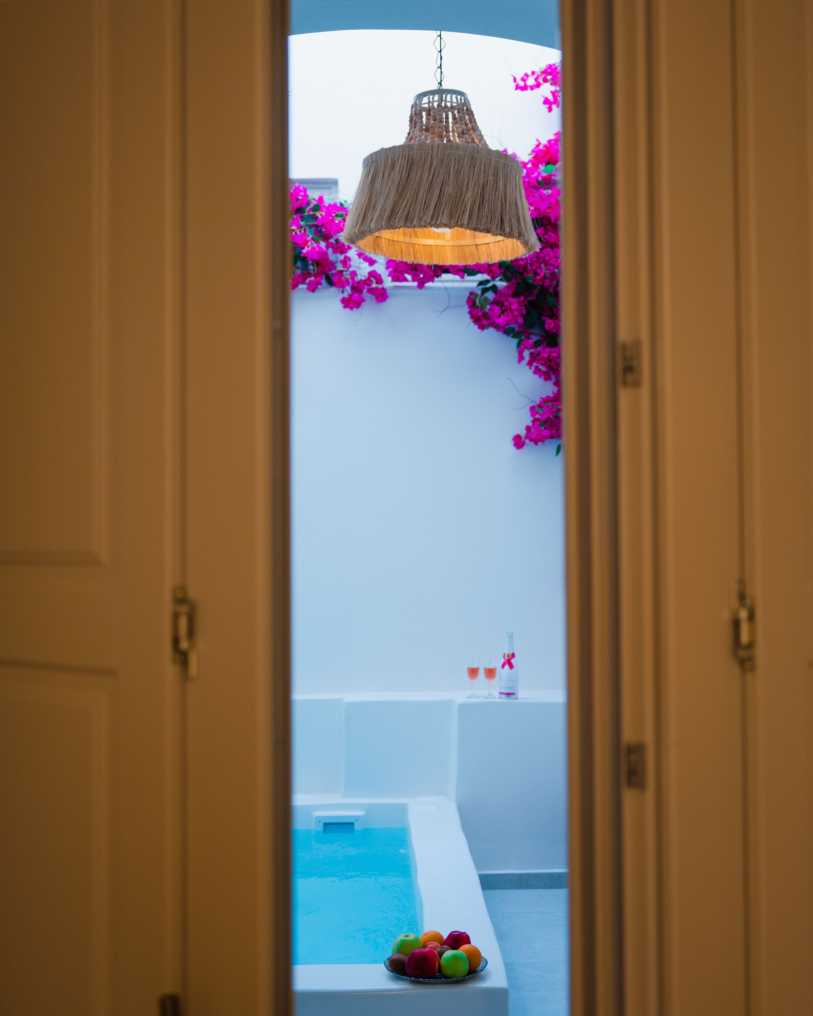 Una puerta abierta que da a una zona de piscina con una lámpara colgante y flores rosadas.