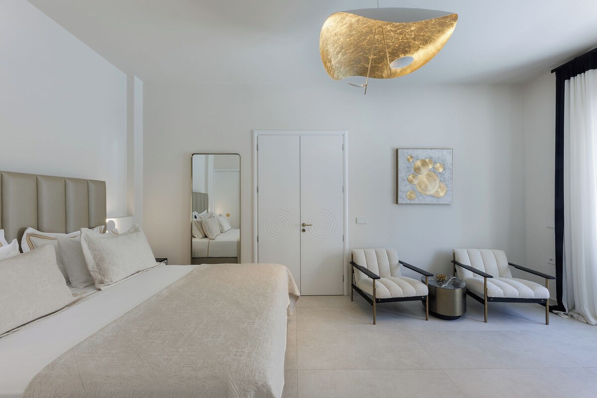 A modern hotel room with a bed, two armchairs, a round coffee table, a large mirror, and a unique ceiling light fixture.