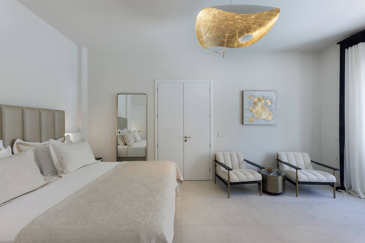 Una habitación de hotel moderna con una cama, dos sillones, una mesa redonda de centro, un espejo grande y una lámpara de techo original.
